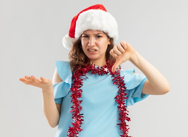 Jonge vrouw in blauwe top en kerstmuts met klatergoud om haar nek kijkt ontevreden en verward met duimen naar beneden arm omhoog in ongenoegen