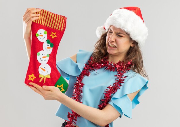 Jonge vrouw in blauwe top en kerstmuts met klatergoud om haar nek met kerstsok te kijken met walging expressie