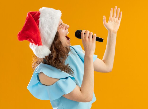 Jonge vrouw in blauwe top en kerstmuts schreeuwen naar microfoon opgewonden gek emotioneel met opgeheven arm