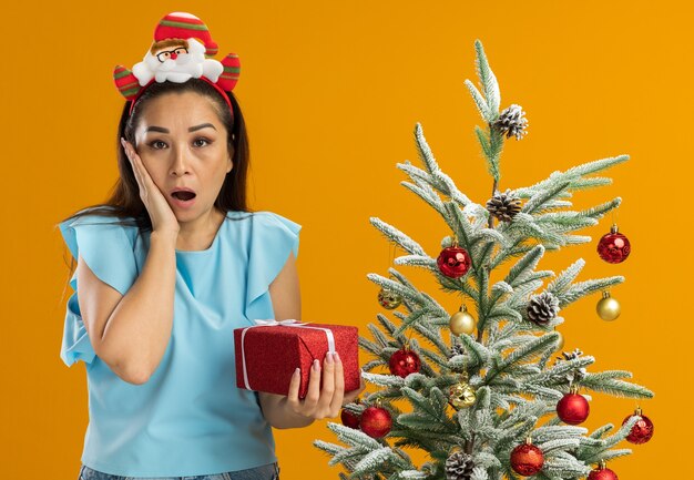 Jonge vrouw in blauwe top met grappige kerstrand op hoofd bedrijf aanwezig camera kijken verbaasd met hand op haar wang staande naast een kerstboom over oranje achtergrond