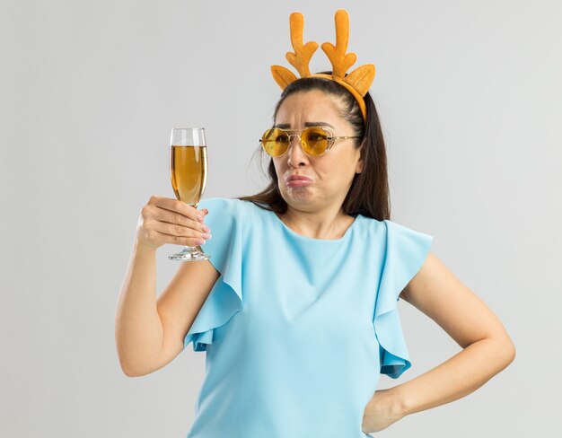Jonge vrouw in blauwe top met grappige rand met herten hoorns en gele glazen met glas champagne kijken verward en ontevreden