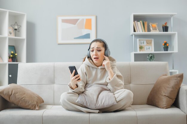 Jonge vrouw in casual kleding met koptelefoon zittend op een bank in het interieur met smartphone die er verward en ontevreden uitziet