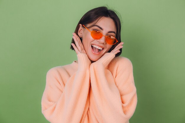 Jonge vrouw in casual perzik en oranje bril trui geïsoleerd op groene olijf muur opgewonden mond open te houden spreidende handen