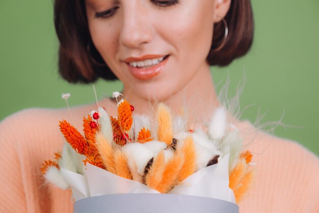 Jonge vrouw in casual perzik trui geïsoleerd op groene olijf muur houden oranje witte bloembak samenstelling van katoen bloemen gypsophila tarwe en lagurus voor een geschenk blij verbaasd verrast