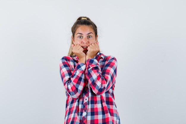 Jonge vrouw in casual shirt met vuisten in de buurt van de mond en bang, vooraanzicht op zoek.