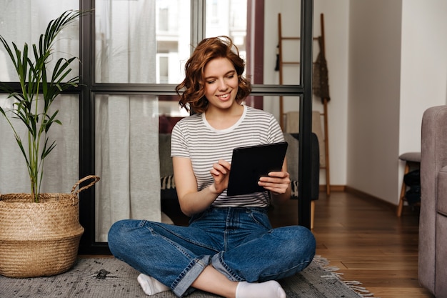 Jonge vrouw in comfortabele kleding met glimlach kijkt naar tablet en vormt in de woonkamer.