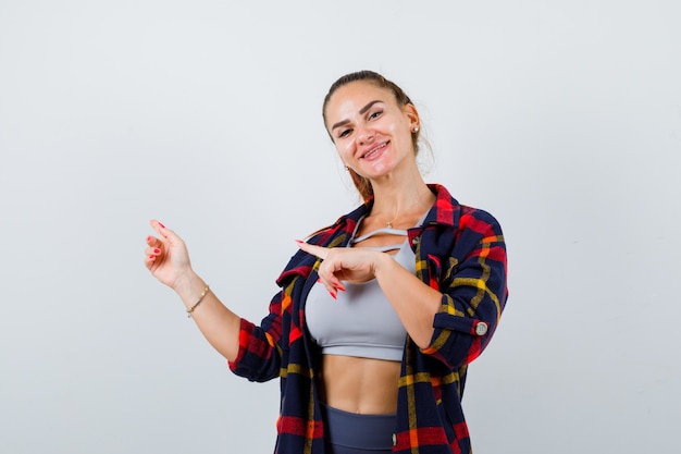 Jonge vrouw in crop top, geruit hemd, broek die naar de linkerkant wijst en er gelukkig uitziet, vooraanzicht.