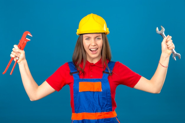 Gratis foto jonge vrouw in de bouw uniforme en gele veiligheidshelm permanent met opgeheven handen met verstelbare sleutels geërgerd en gefrustreerd schreeuwen met woede op blauw geïsoleerde achtergrond