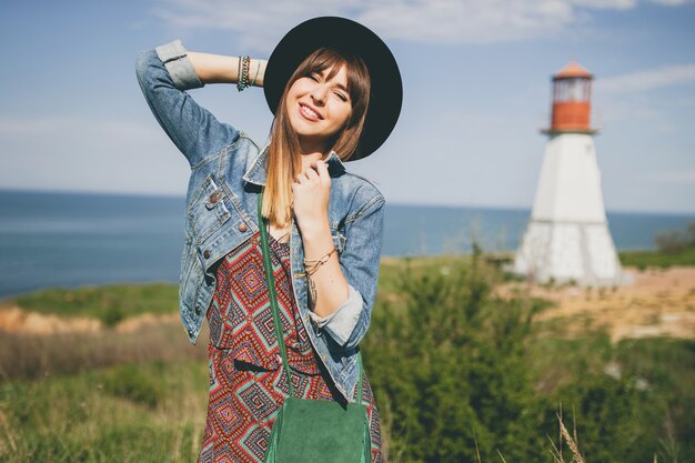 Jonge vrouw in de natuur, vuurtoren, bohemien outfit, spijkerjasje, zwarte hoed, glimlachen, gelukkig, zomer, stijlvolle accessoires