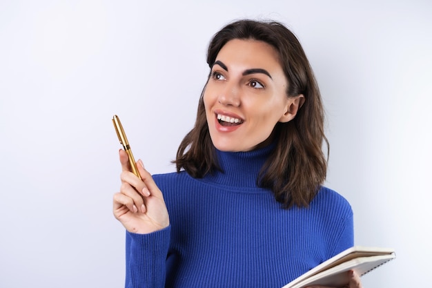 Jonge vrouw in een blauwe golf coltrui op een witte achtergrond peinzend met een notitieboekje in haar hand denkt over ideeën doelen voor het jaar