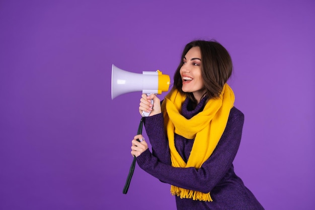 Jonge vrouw in een gebreide jurk en in een gele sjaal op een paarse achtergrond schreeuwt vrolijk in een megafoon, vrolijk, kondigt een verkoop aan