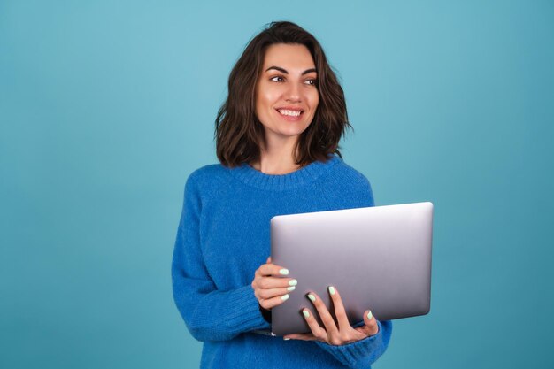 Jonge vrouw in een geïsoleerde gebreide trui houdt een laptop vast, kijkt naar het scherm en lacht vrolijk