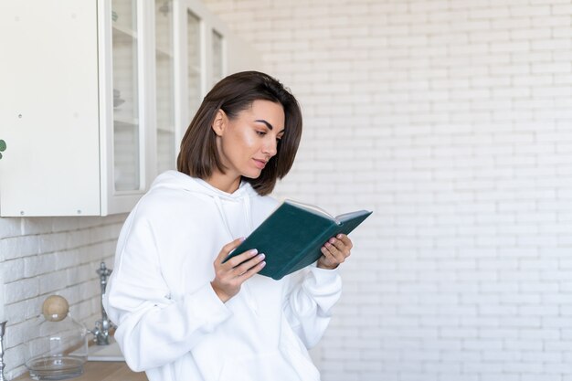 Jonge vrouw in een warme witte hoodie thuis in de keuken leest 's ochtends een boek