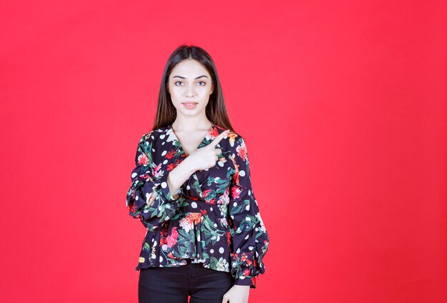 Jonge vrouw in gebloemd hemd dat op de rode muur staat en naar rechts wijst