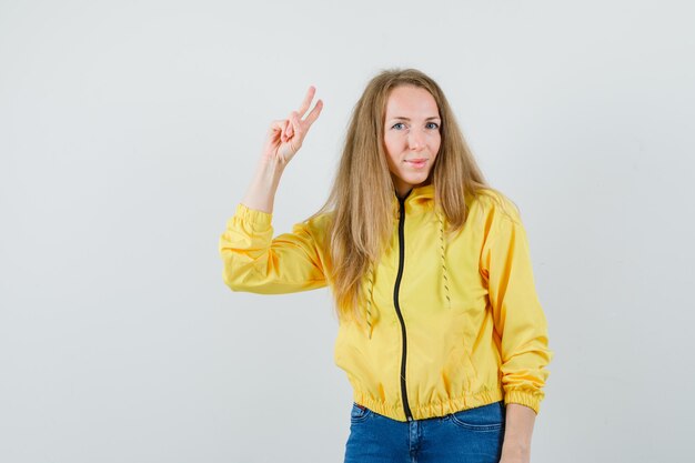 Jonge vrouw in geel bomberjack en blauwe jean met pistoolgebaar en op zoek naar optimistisch, vooraanzicht.