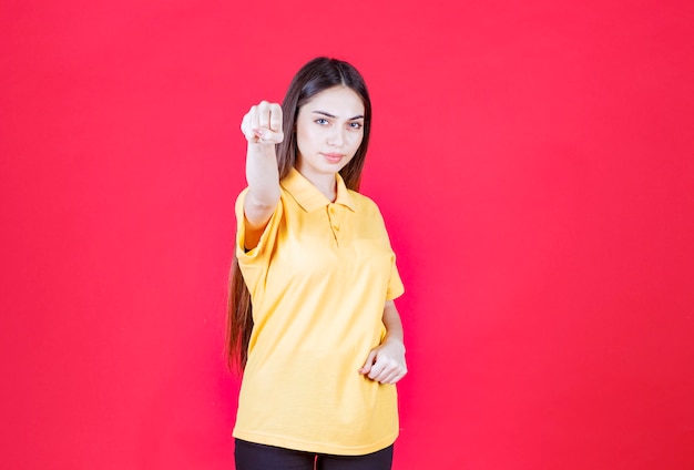 Jonge vrouw in geel overhemd die zich op rode muur bevindt en positief handteken toont