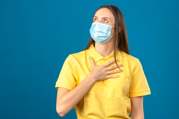 Jonge vrouw in geel poloshirt en medisch beschermend masker die omhooggaand en wat betreft haar borst op blauw geïsoleerde achtergrond kijken