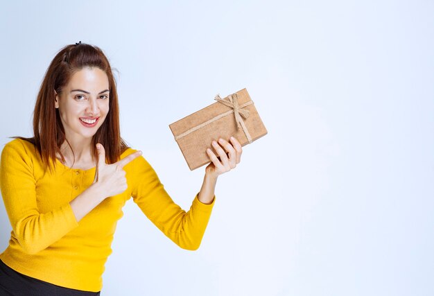 Jonge vrouw in geel shirt die een kartonnen geschenkdoos vasthoudt en een positief handteken toont