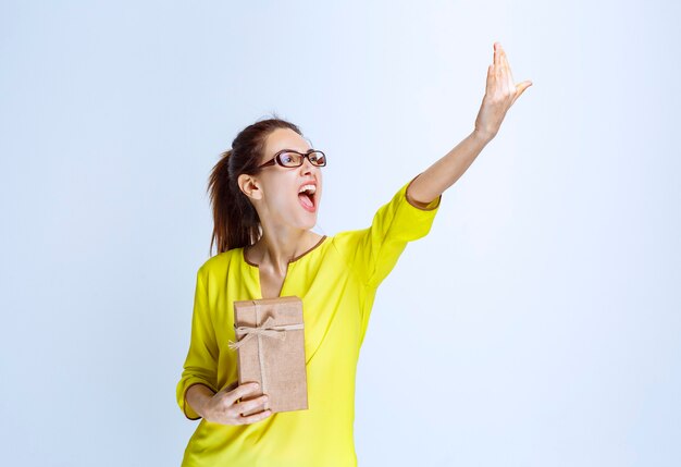 Jonge vrouw in geel shirt die een kartonnen geschenkdoos vasthoudt en iemand uitnodigt om het te presenteren