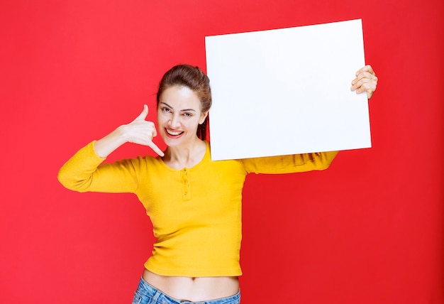 Jonge vrouw in geel shirt die een vierkant infobord vasthoudt en om een telefoontje vraagt