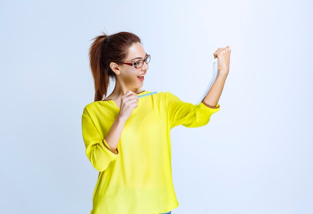 Jonge vrouw in geel shirt die haar examenblad vasthoudt en denkt terwijl ze een pen vasthoudt