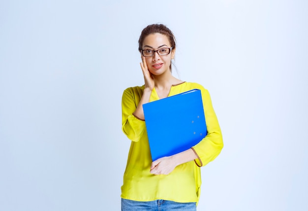 Jonge vrouw in geel shirt met een blauwe map en kijkt verward en verrast