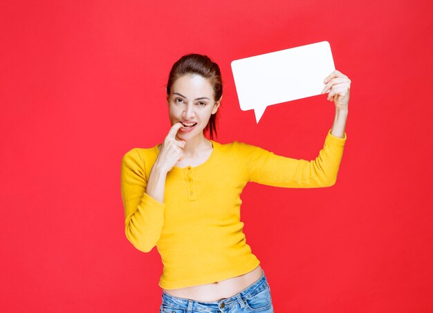 Jonge vrouw in geel shirt met een rechthoekig infobord en ziet er verward en onzeker uit