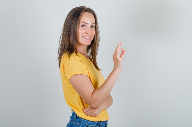 Jonge vrouw in geel t-shirt, korte broek die ok teken doet en vrolijk kijkt.