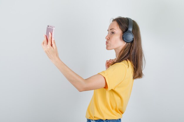 Jonge vrouw in geel t-shirt, korte broek praten over smartphone via videocall.