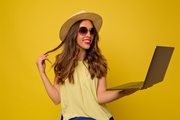 Jonge vrouw in gele jurk met hoed en zonnebril luisteren naar muziek met behulp van laptop