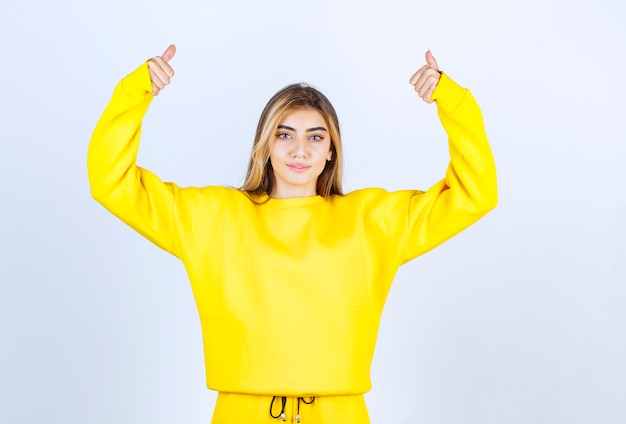 Jonge vrouw in gele sweatsuit die duimen opgeeft op witte muur