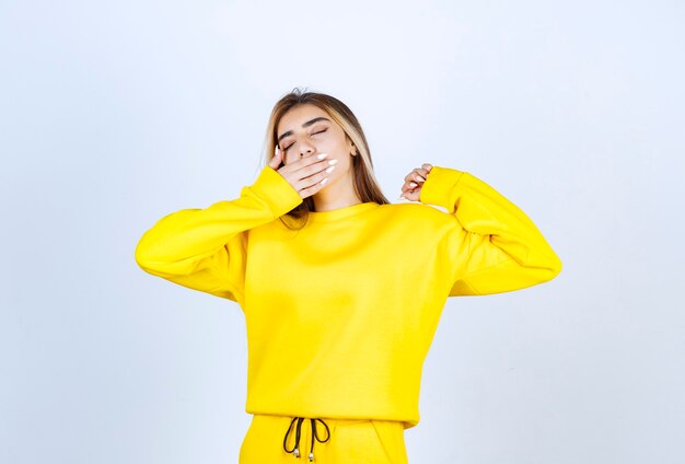 Jonge vrouw in gele sweatsuit die zich slaperig voelt over witte muur