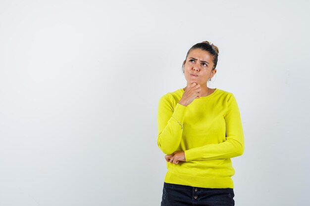 Jonge vrouw in gele trui en zwarte broek die in denkende pose staat en peinzend kijkt