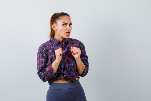 Jonge vrouw in geruit hemd, broek met handen over de borst en verbaasd, vooraanzicht.