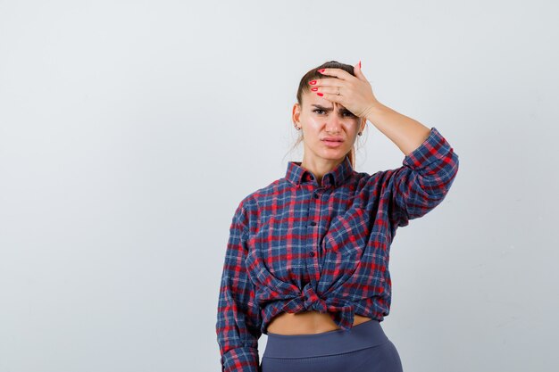 Jonge vrouw in geruit hemd die hoofd met palm vasthoudt en er pijnlijk uitziet, vooraanzicht.