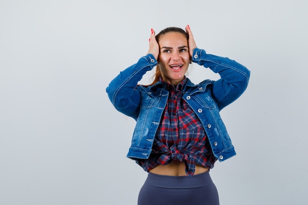 Jonge vrouw in geruit hemd, jas, broek met handen op het hoofd en vrolijk, vooraanzicht.