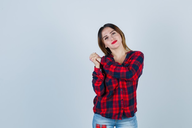 Jonge vrouw in geruit hemd poseren terwijl ze de handen omklemt, het hoofd op de schouder buigt en beschaamd kijkt, vooraanzicht.