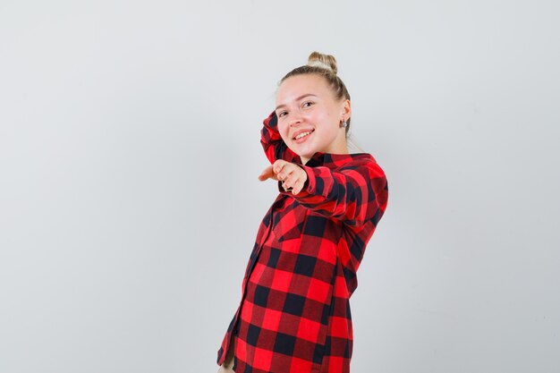 Gratis foto jonge vrouw in geruit overhemd dat zich met uitgestrekte hand bevindt en dartel kijkt