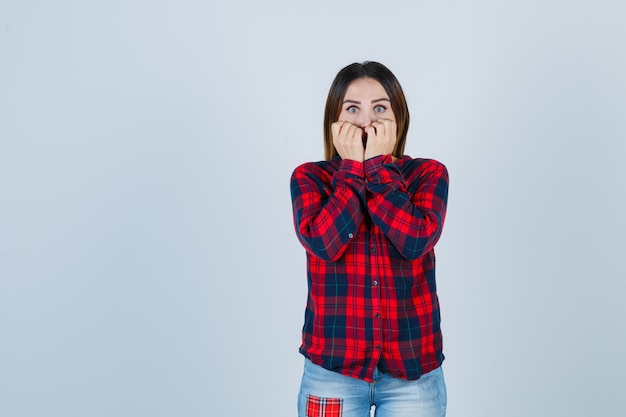 Jonge vrouw in geruit overhemd, jeans die emotioneel vingers bijt en angstig kijkt, vooraanzicht.