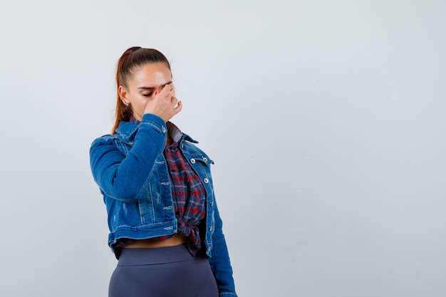 Jonge vrouw in geruit overhemd, spijkerjasje met hand op gezicht, ogen sluiten en er moe uitzien.