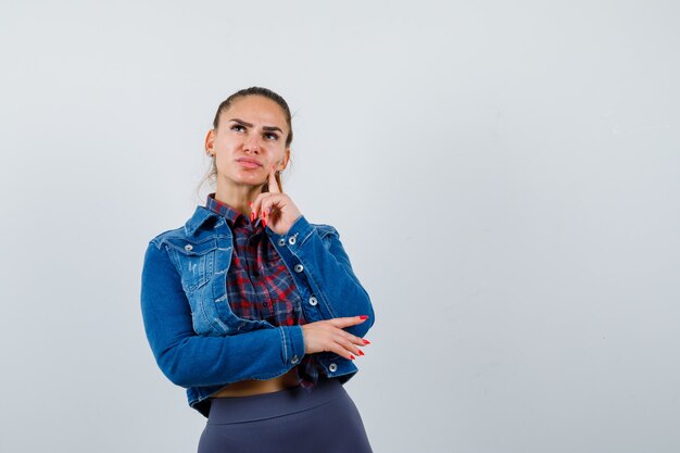 Jonge vrouw in geruit overhemd, spijkerjasje staat in denkende pose en ziet er verstandig uit, vooraanzicht.