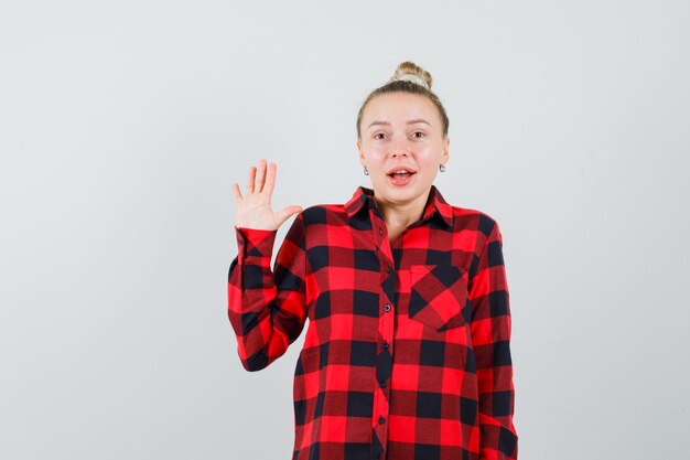 Jonge vrouw in geruit overhemd zwaaiende hand voor groet en op zoek verbaasd, vooraanzicht.