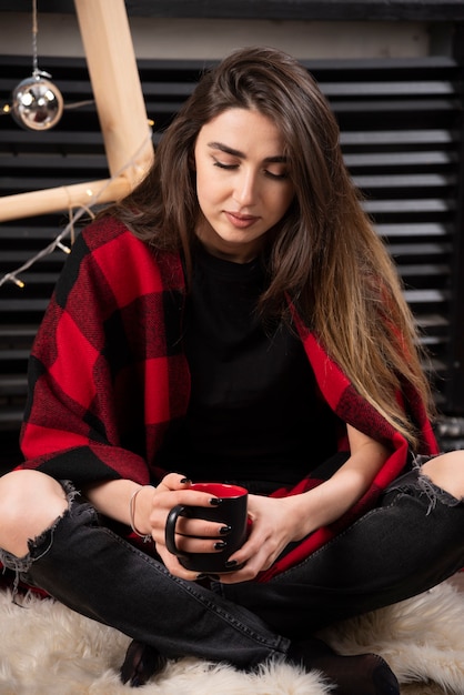 Jonge vrouw in geruite plaid zittend op de vloer en met een kopje.