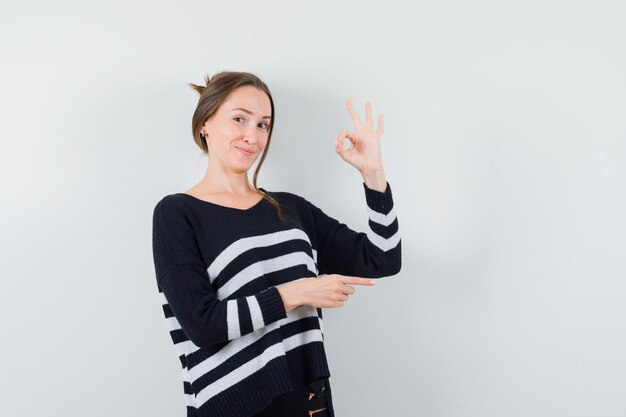 Jonge vrouw in gestreept breigoed en zwarte broek die ok teken tonen en naar rechts wijzen en gelukkig kijken