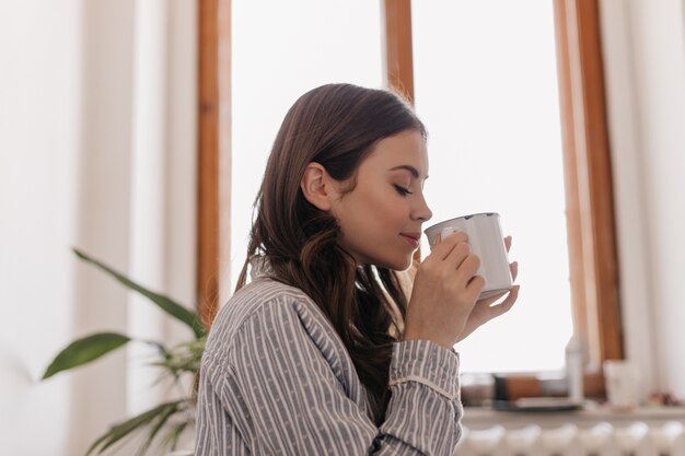 Jonge vrouw in gestreept overhemd drinkt koffie uit ijzeren beker tegen raam