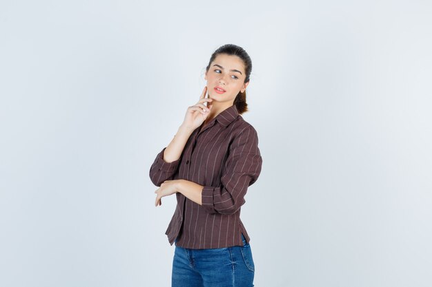 Jonge vrouw in gestreept shirt, jeans die wijsvinger op de kaak legt, wegkijkt terwijl ze poseert en er charmant uitziet, vooraanzicht.