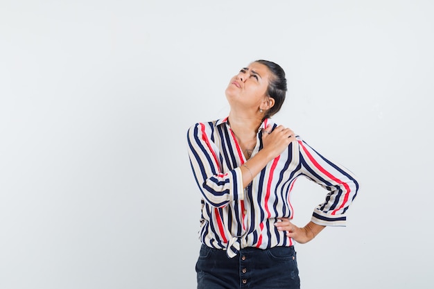 Jonge vrouw in gestreepte blouse die één hand op taille, een andere hand op schouder legt en over iets denkt en peinzend kijkt