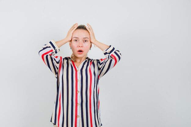 Jonge vrouw in gestreepte blouse die haar hoofd bedekt met beide handen en geschokt kijkt