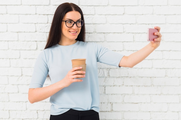 Jonge vrouw in glazen die selfie met koffiekop nemen