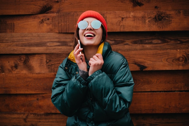 Gratis foto jonge vrouw in goed humeur lacht oprecht tijdens het praten over de telefoon meisje in rode hoed en emerald kleur jas poses op houten achtergrond